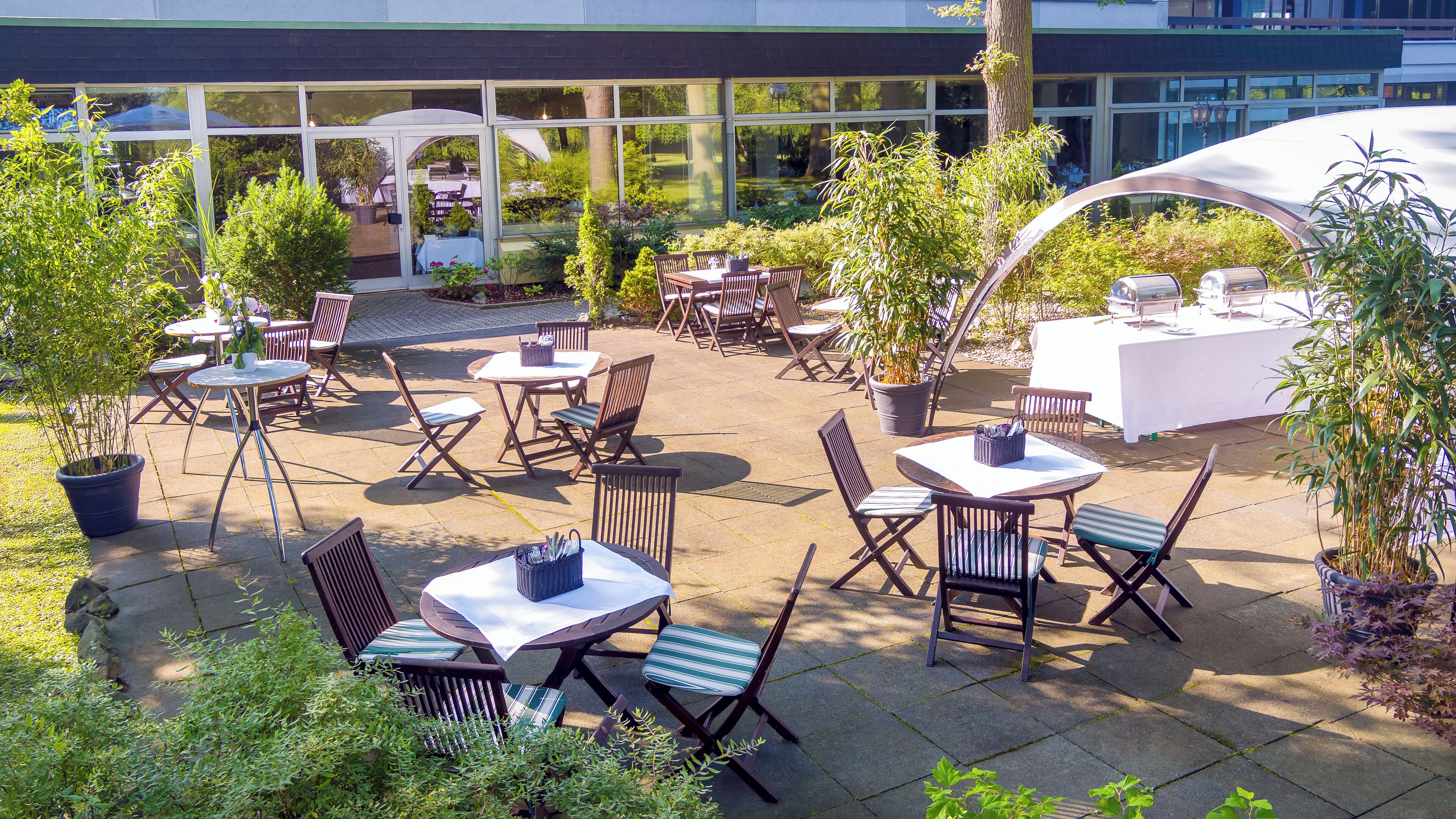 Een terras buiten met meerdere tafels en stoelen, omgeven door potplanten en bomen.