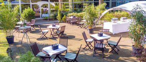 An outdoor patio area with multiple tables and chairs, surrounded by potted plants and trees.