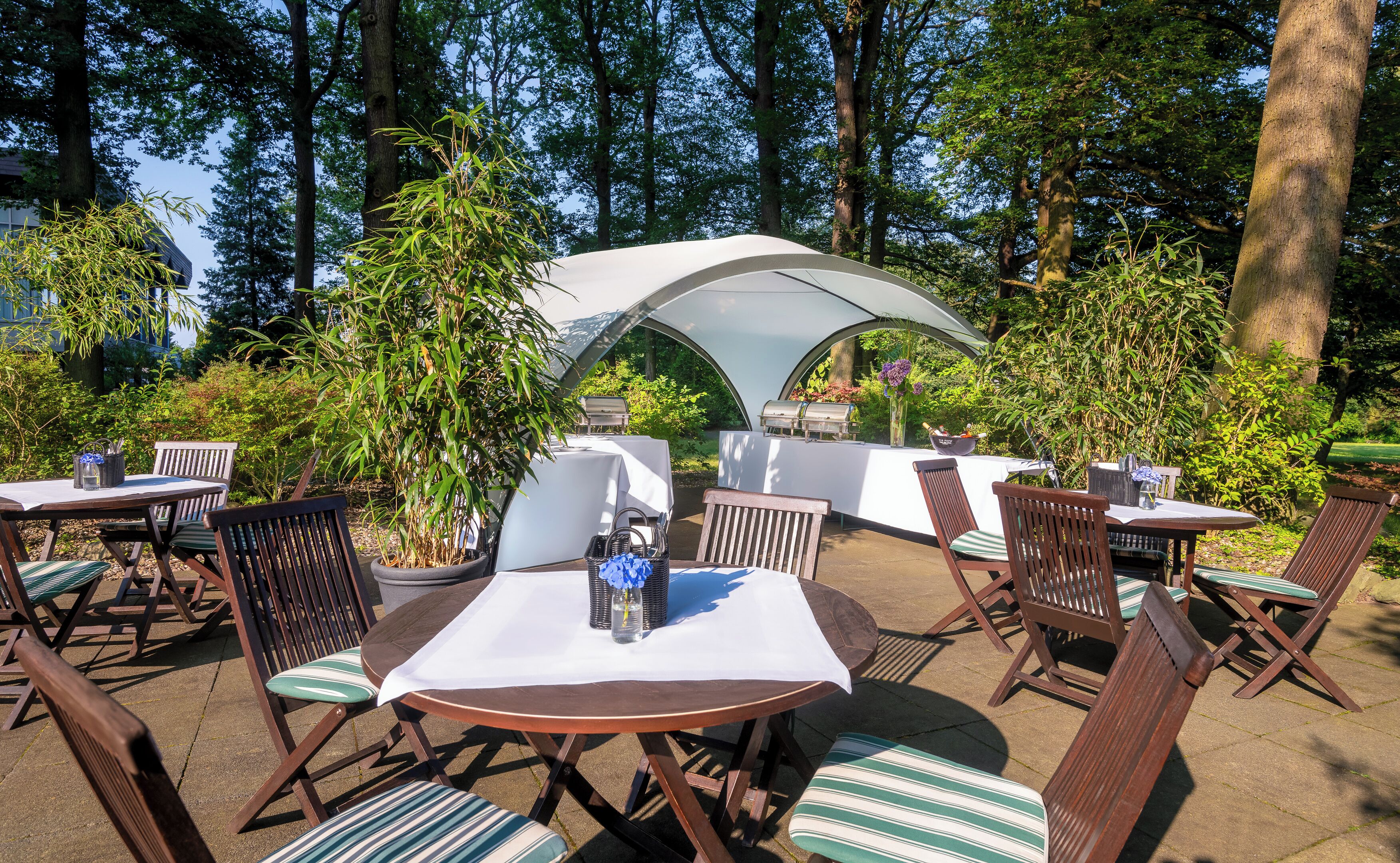 En udendørs terrasse med runde borde og stole, overdækket af et hvidt markise, omgivet af grønt og træer.