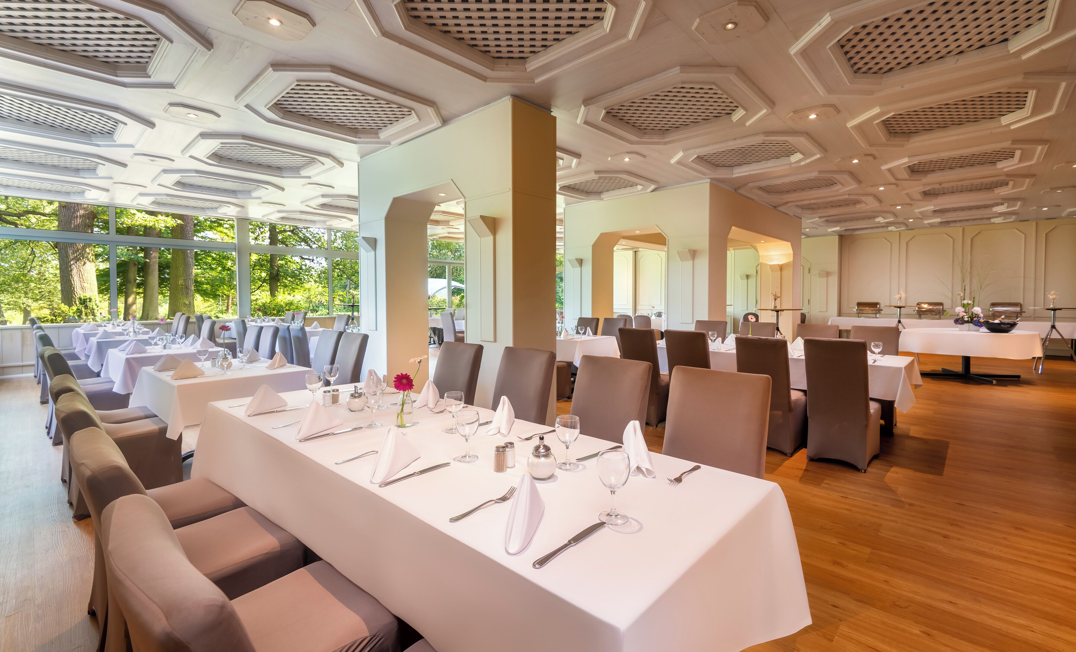 A spacious dining area with multiple tables set for service, large windows offering a view of greenery, and a high ceiling with geometric patterns.