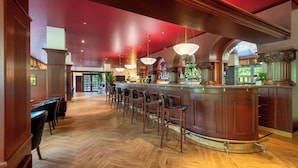 Zona de bar con suelo de madera, varios taburetes altos y una barra con gran variedad de botellas.