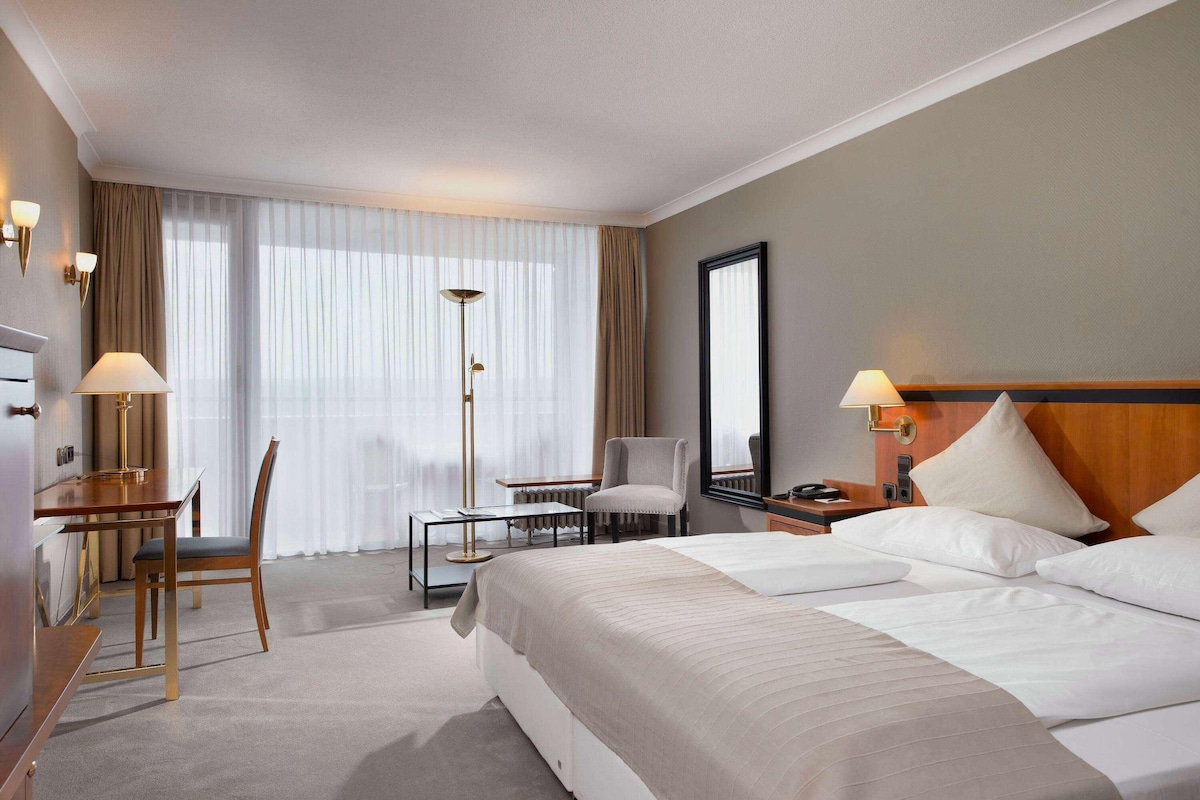 A hotel room with a large bed, a desk with a chair, a standing lamp, and a large window with curtains.