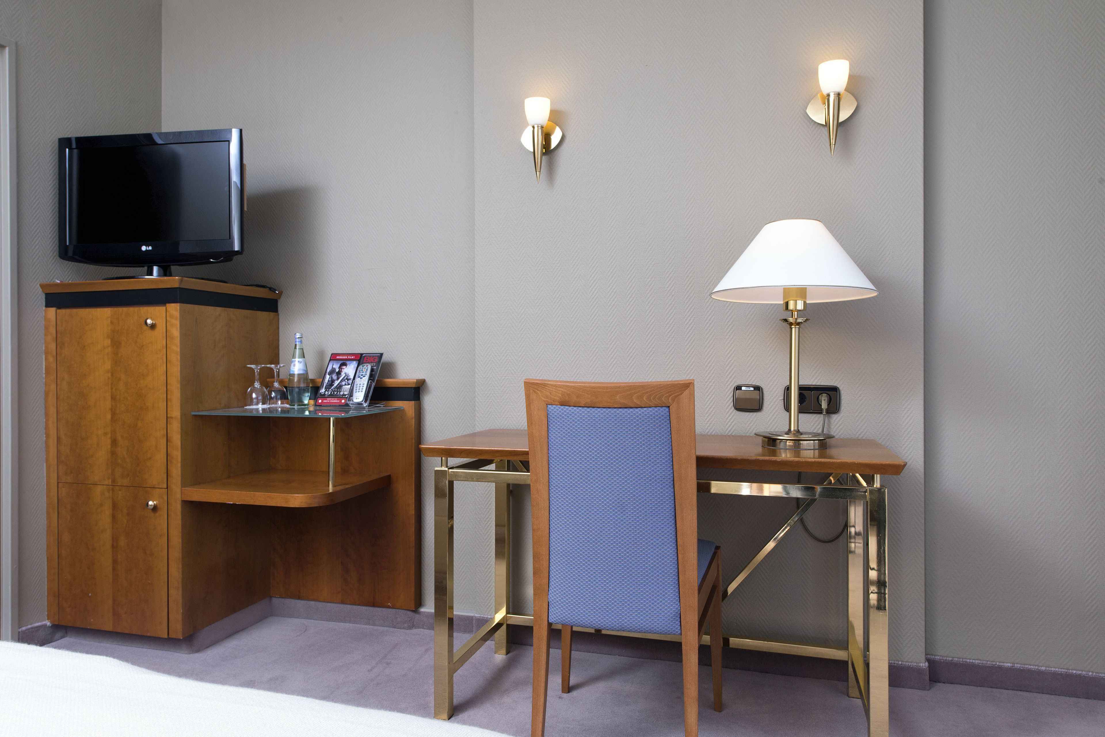 Quarto de hotel com televisão sobre um móvel de madeira, uma escrivaninha com abajur e uma cadeira.
