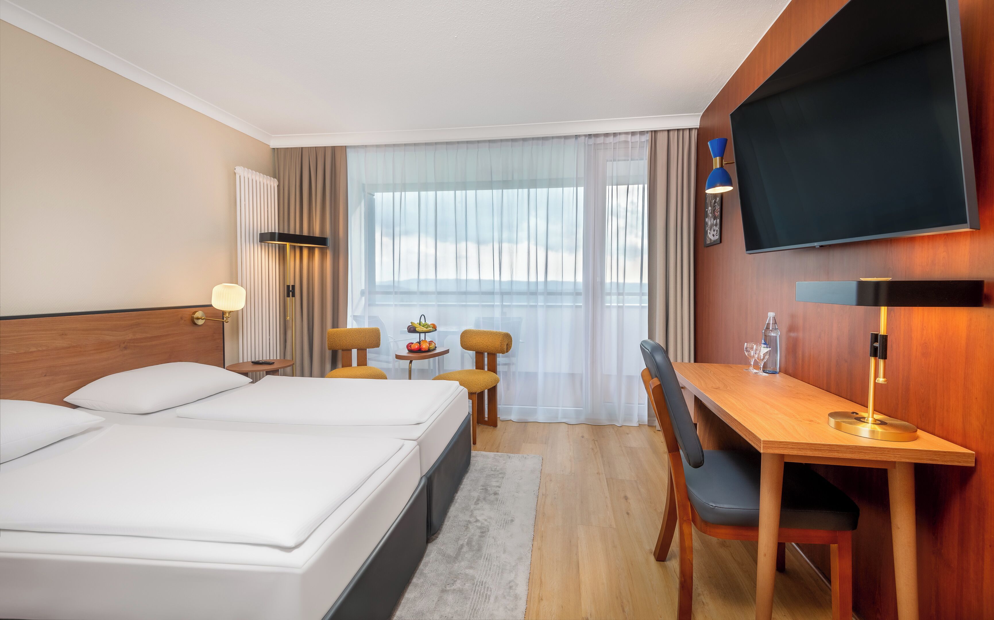 A hotel room with a large bed, a desk with a chair, a television, and a view of the outdoors through the window.