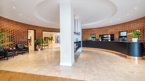 Un área de recepción moderna con un mostrador negro curvo, pilares blancos y una pared con diseño. Hay zonas de estar con sillas y plantas en macetas.