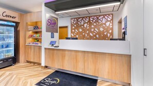 Un espace d’accueil d’hôtel comprenant un comptoir de réception en bois, un réfrigérateur rempli de boissons et un paillasson.