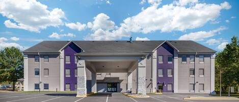 A multi-story hotel with a covered entrance, parking lot, and a clear blue sky with clouds.