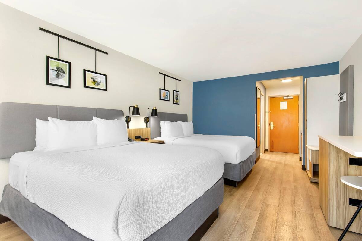 Une chambre d’hôtel avec deux lits, un bureau et une chaise. Des tableaux encadrés ornent le mur, et une porte donne accès à une autre pièce.