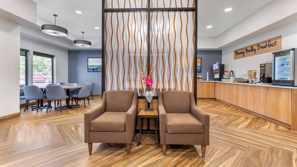 Un espace d’accueil moderne comprenant un comptoir de réception en bois, deux fauteuils marron, un coin café et un espace repas avec des chaises grises et une table.