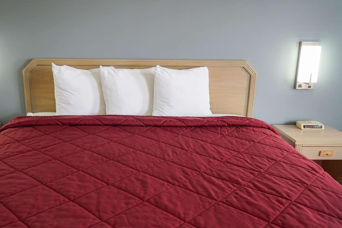 A hotel room with a large bed, a wooden headboard, white pillows, a red quilted bedspread, bedside tables with lamps, a phone, and a digital clock.