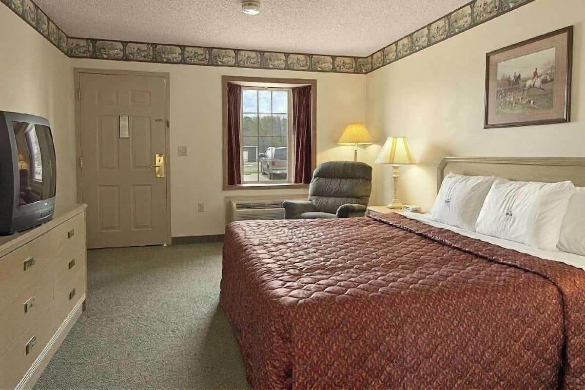 A hotel room with a bed, a chair, a desk, a television, a window, and a painting on the wall.