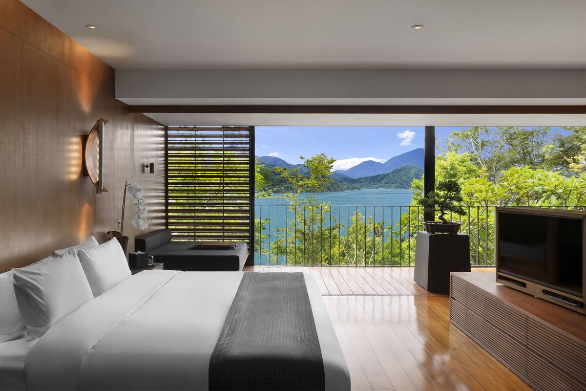 A modern bedroom with a large bed, a TV, and a view of a lake and mountains.