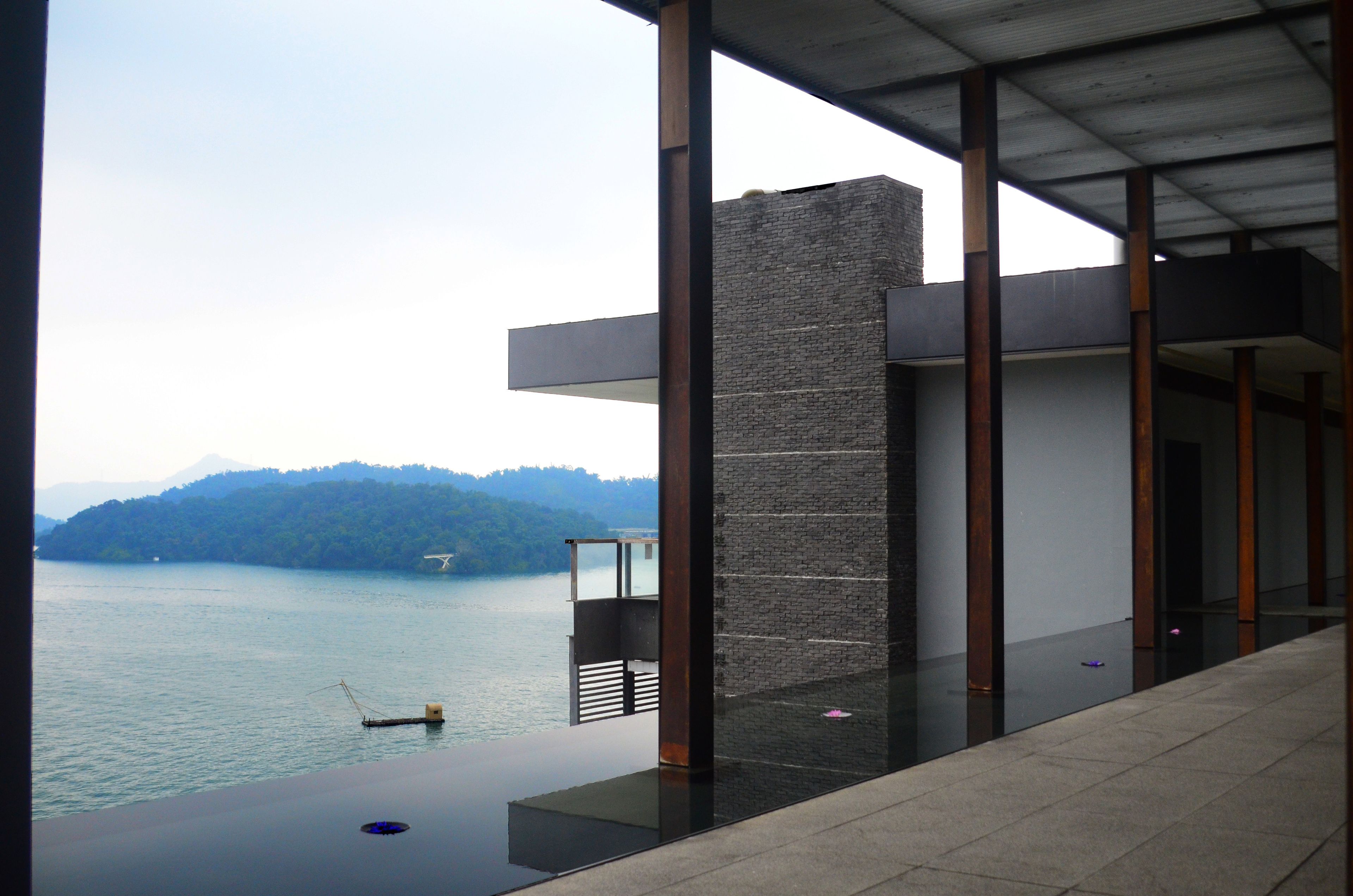 A modern building with large glass windows overlooking a body of water and a forested hill.