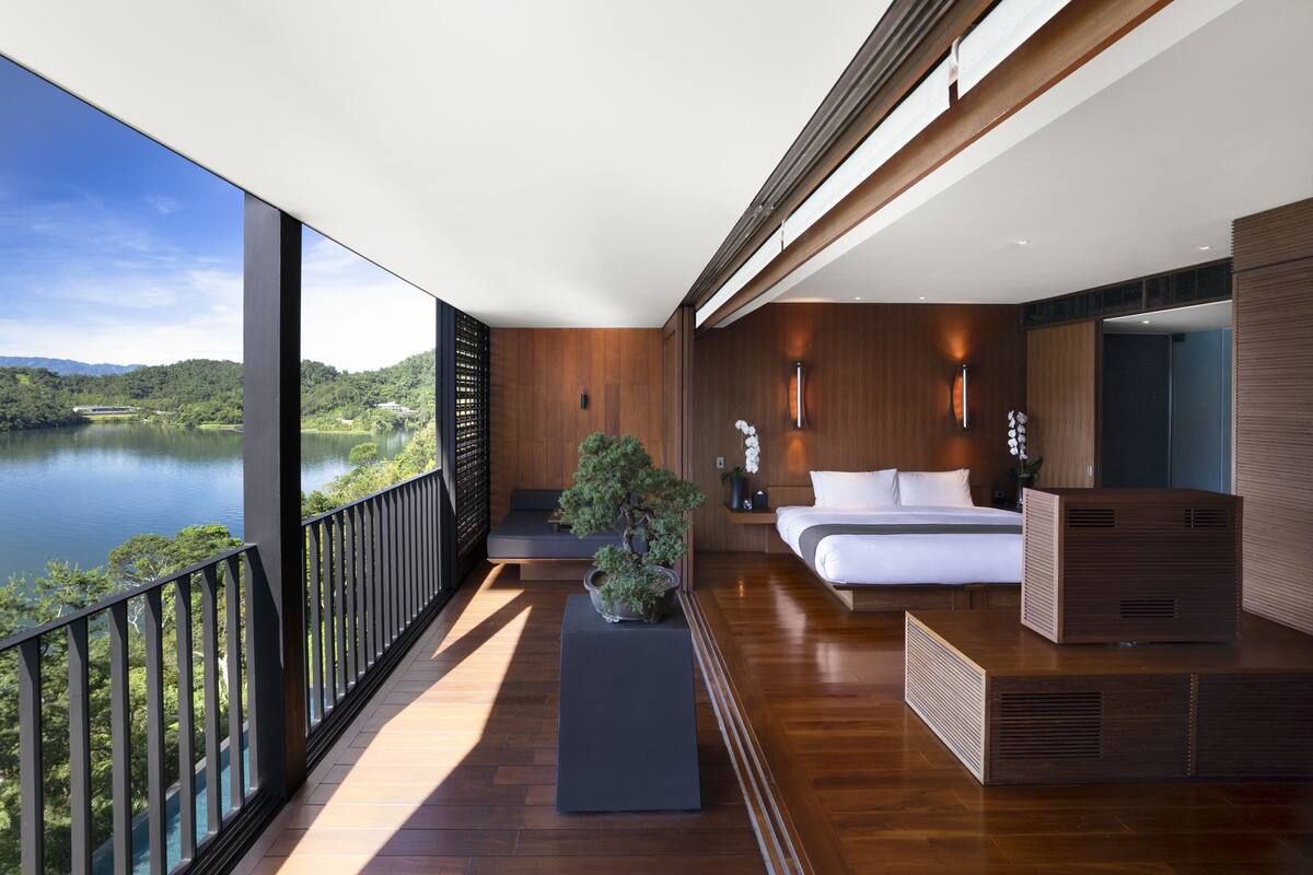 A modern hotel room with a balcony, wooden flooring, a bed, a small seating area, and a potted plant.