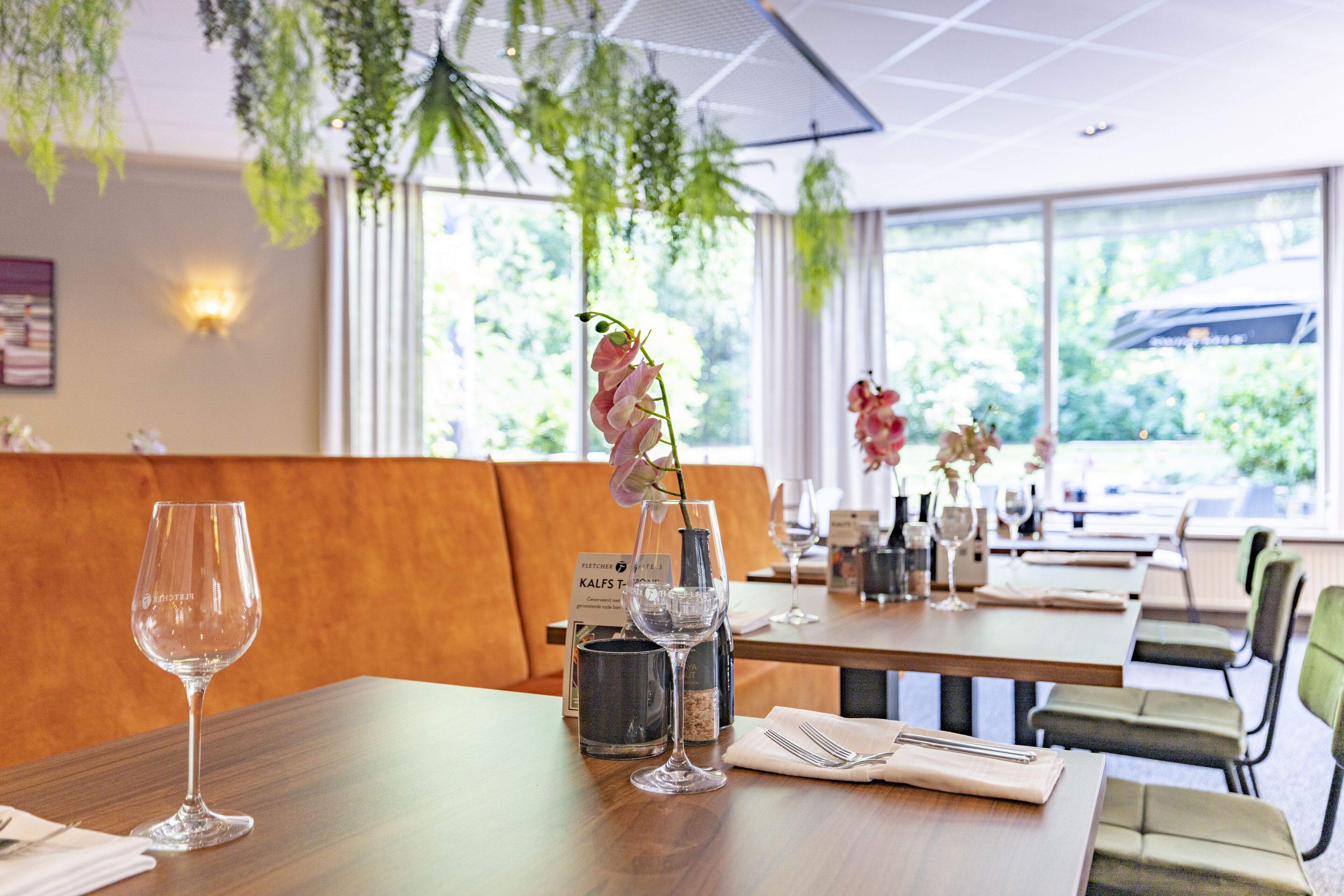 A restaurant with wooden tables, green upholstered chairs, and hanging plants.