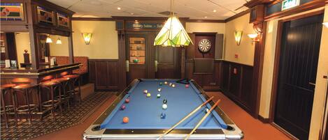 Une table de billard avec des billes et des queues, un jeu de fléchettes, un coin bar avec des tabourets et une enseigne indiquant un salon de beauté.