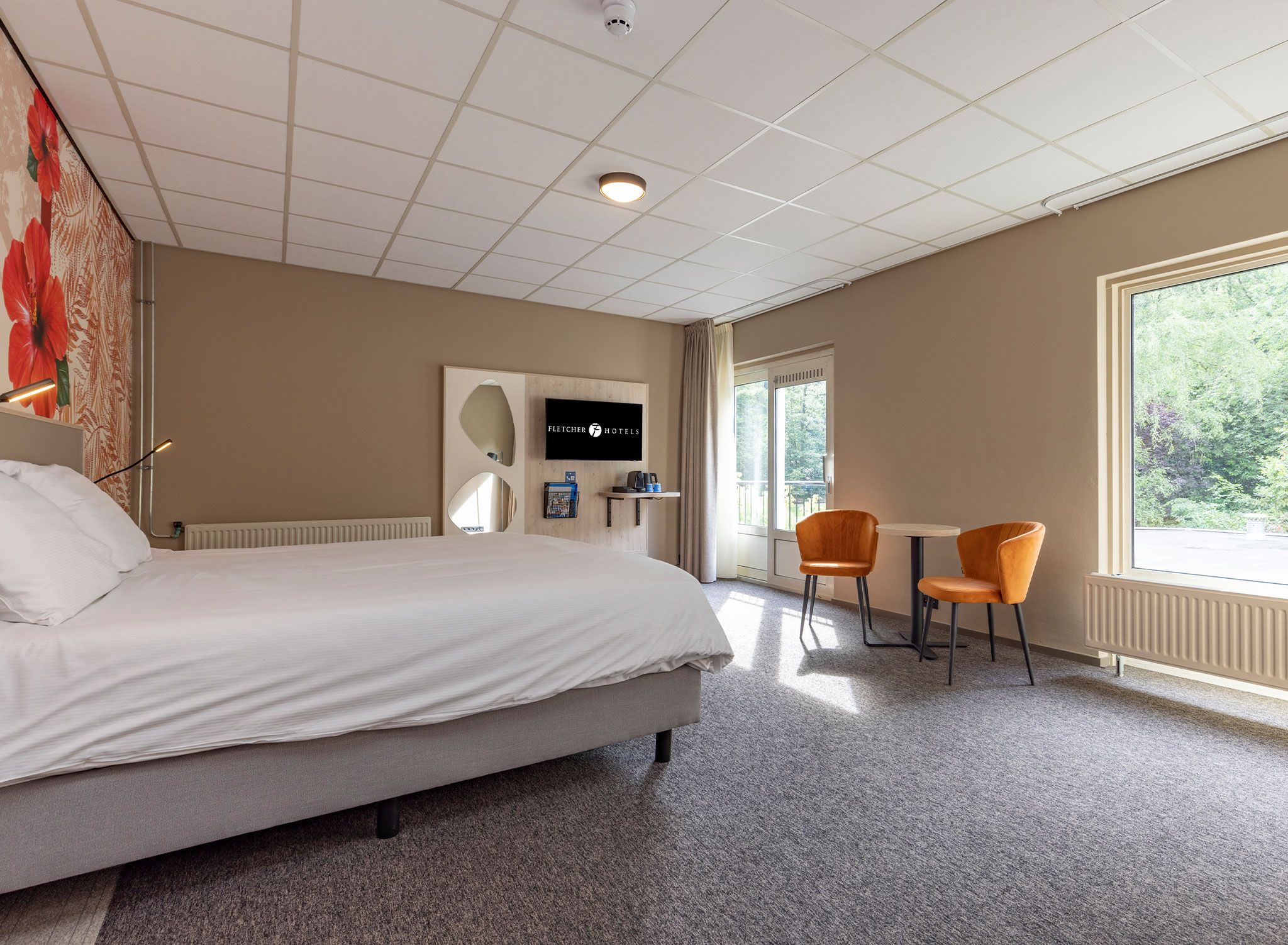 A hotel room with a bed, a seating area with two chairs, a coffee machine, and a view of greenery outside.
