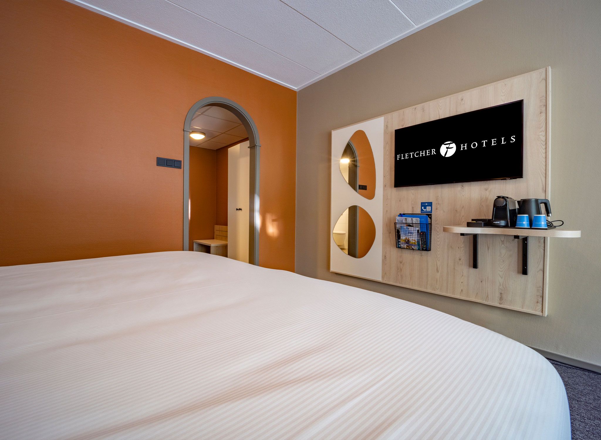 Habitación de hotel con una cama grande, televisión montada y una cafetera en un estante.