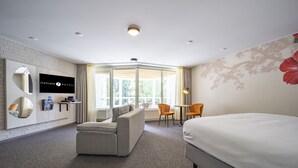 Ein Hotelzimmer mit einem großen Bett, einer Couch, einem an der Wand befestigten Fernseher und Blick ins Freie durch die Fenster.