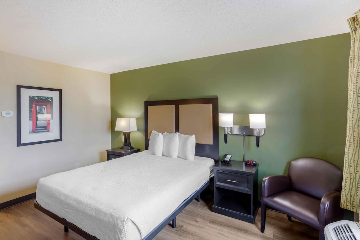 A hotel room with a bed, bedside lamps, a chair, a desk with a phone, and a framed picture on the wall.