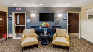 A hotel lobby with two leather armchairs, a round table with a vase of flowers, a flat-screen TV mounted on the wall, and two exit signs. - Extended Stay America Suites New York City LaGuardia Airport (Whitestone)
