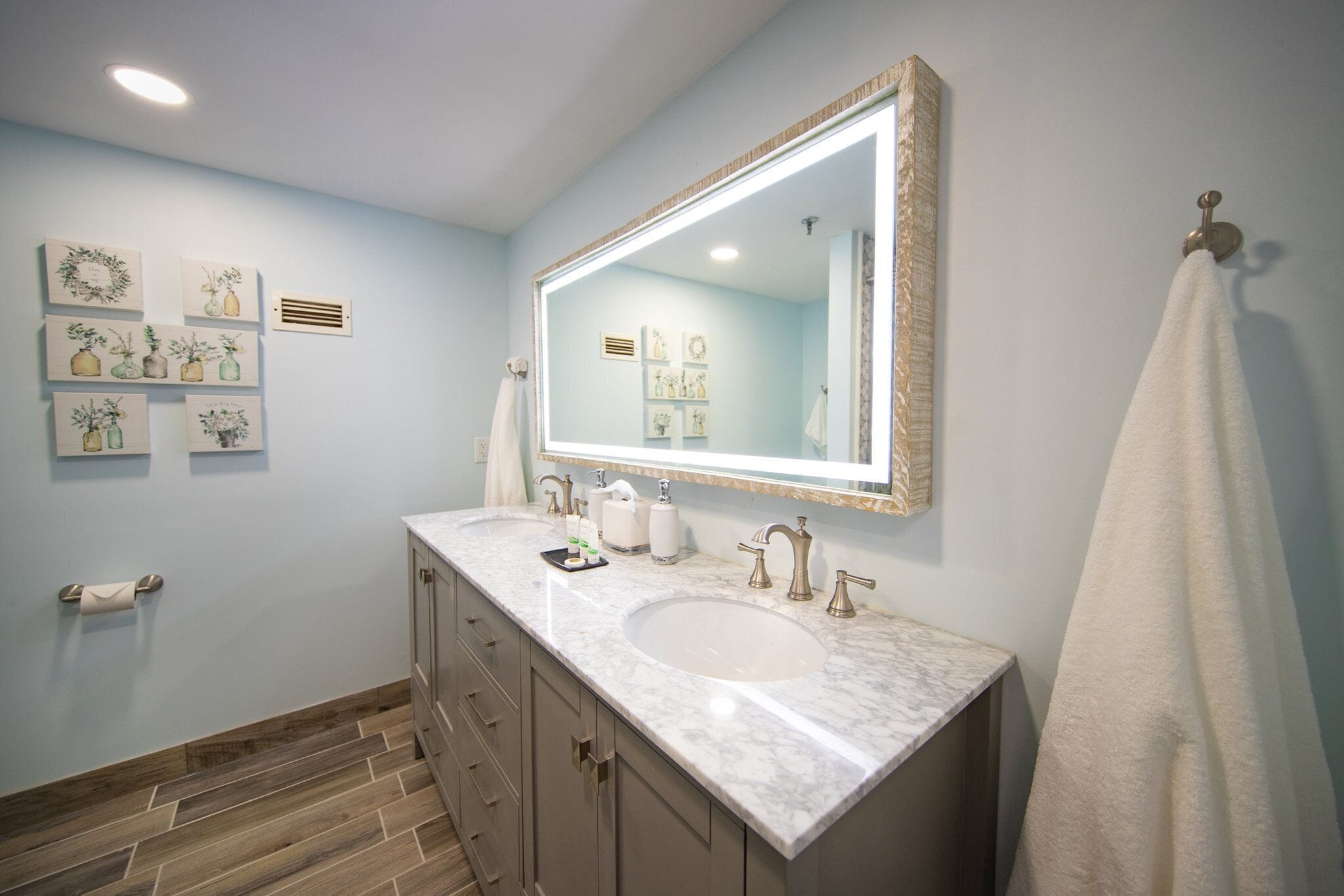 A bathroom with a large mirror, a sink, and a toilet. There are framed pictures on the wall and a towel hanging on the wall.