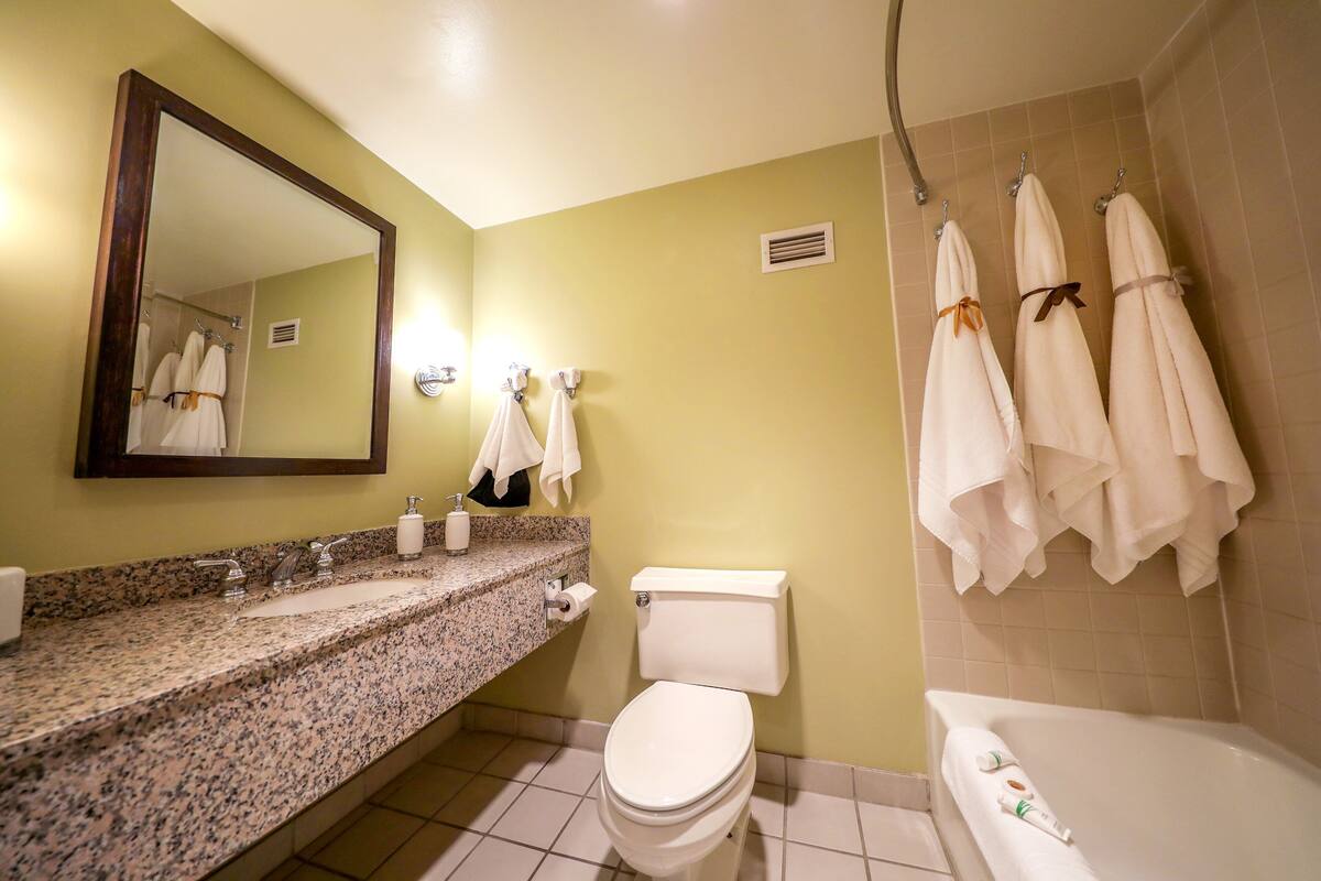 A bathroom with a granite countertop, a white toilet, a bathtub, and multiple white towels hanging on hooks.