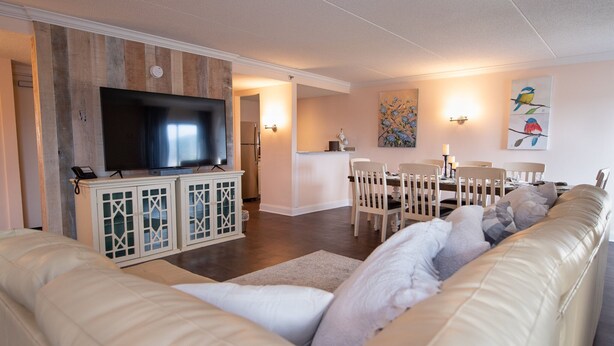 A modern living room with a large flat-screen TV, a beige leather sofa, a dining area with wooden chairs, and a kitchen in the background. - Bella Vista Hotel & Suites (Lynchburg)
