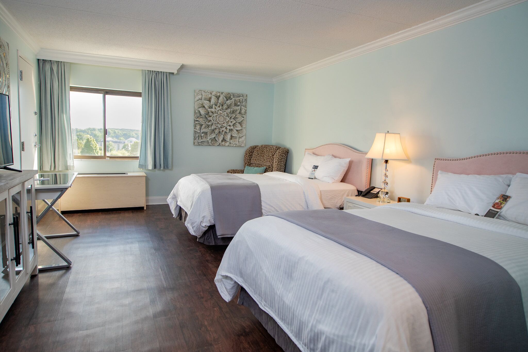 A hotel room with two beds, a desk, a chair, and a large window with curtains.