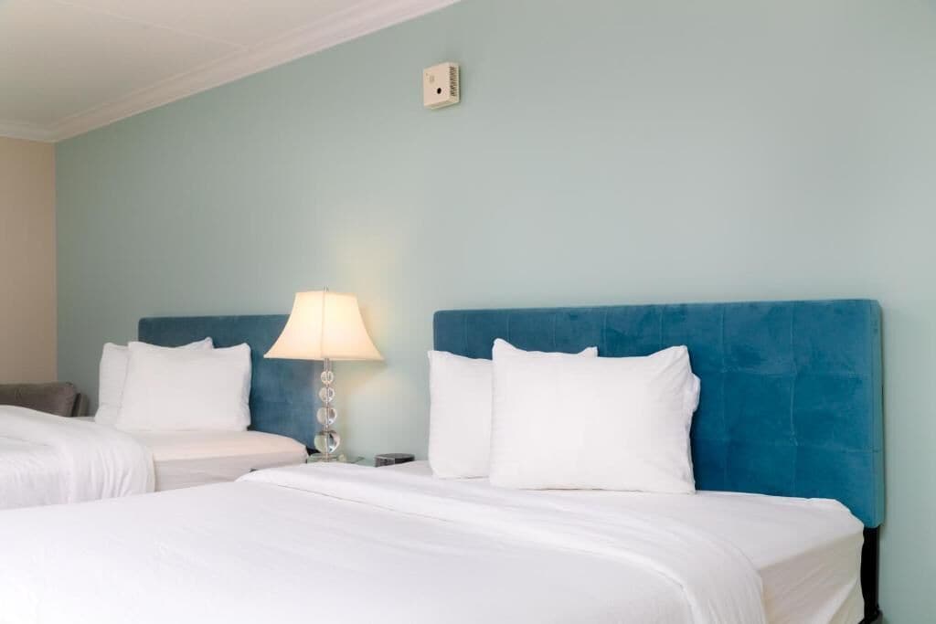 Habitación de hotel con dos camas, cabecera azul, ropa de cama blanca, una lámpara y un interruptor de luz en la pared.