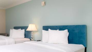 A hotel room with two beds, a blue headboard, white bedding, a lamp, and a wall-mounted light switch.