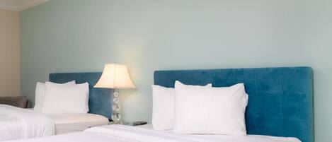 Habitación de hotel con dos camas, cabecera azul, ropa de cama blanca, una lámpara y un interruptor de luz en la pared.