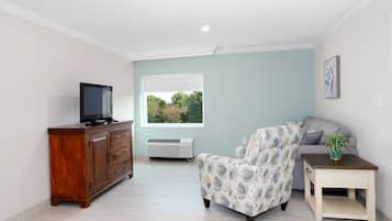 A living room with a wooden cabinet, a television, a floral-patterned armchair, a grey sofa, a small table with a plant, and a window with a view of trees.