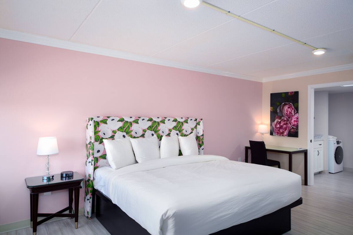 A bedroom with a bed, bedside tables, a desk, a chair, a lamp, and a washing machine.