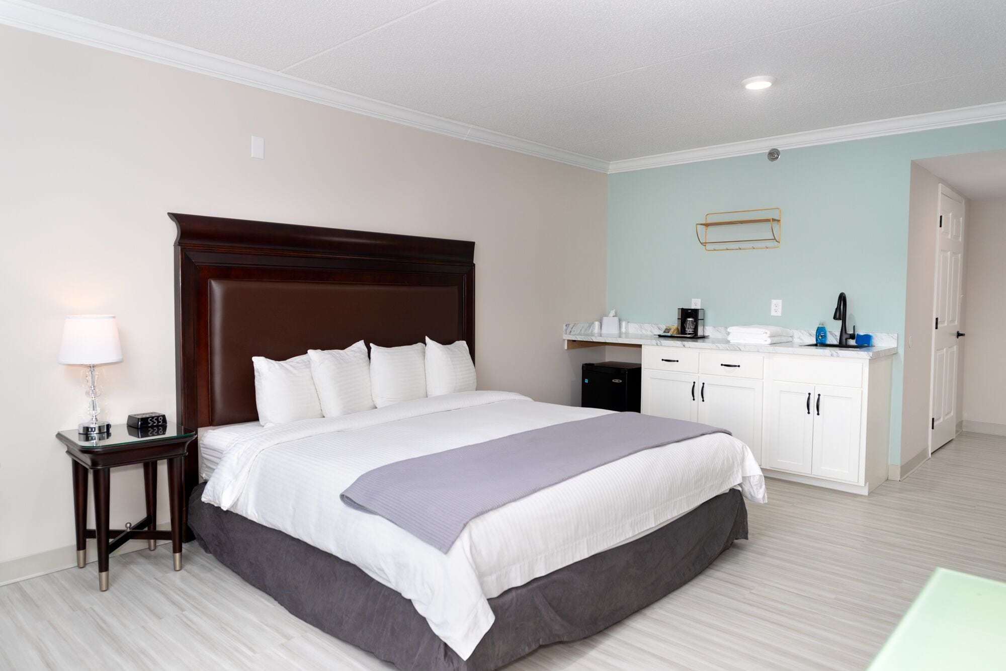 Habitación de hotel con una cama grande, una mesita de noche con lámpara, un escritorio pequeño con cafetera y un baño visible a través de una puerta abierta.