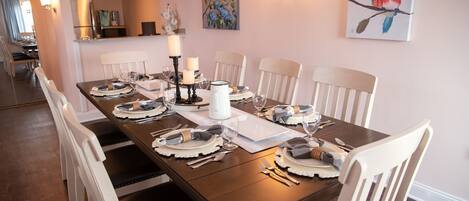 A dining area with a long table set for six, white chairs, and a painting of birds on the wall.