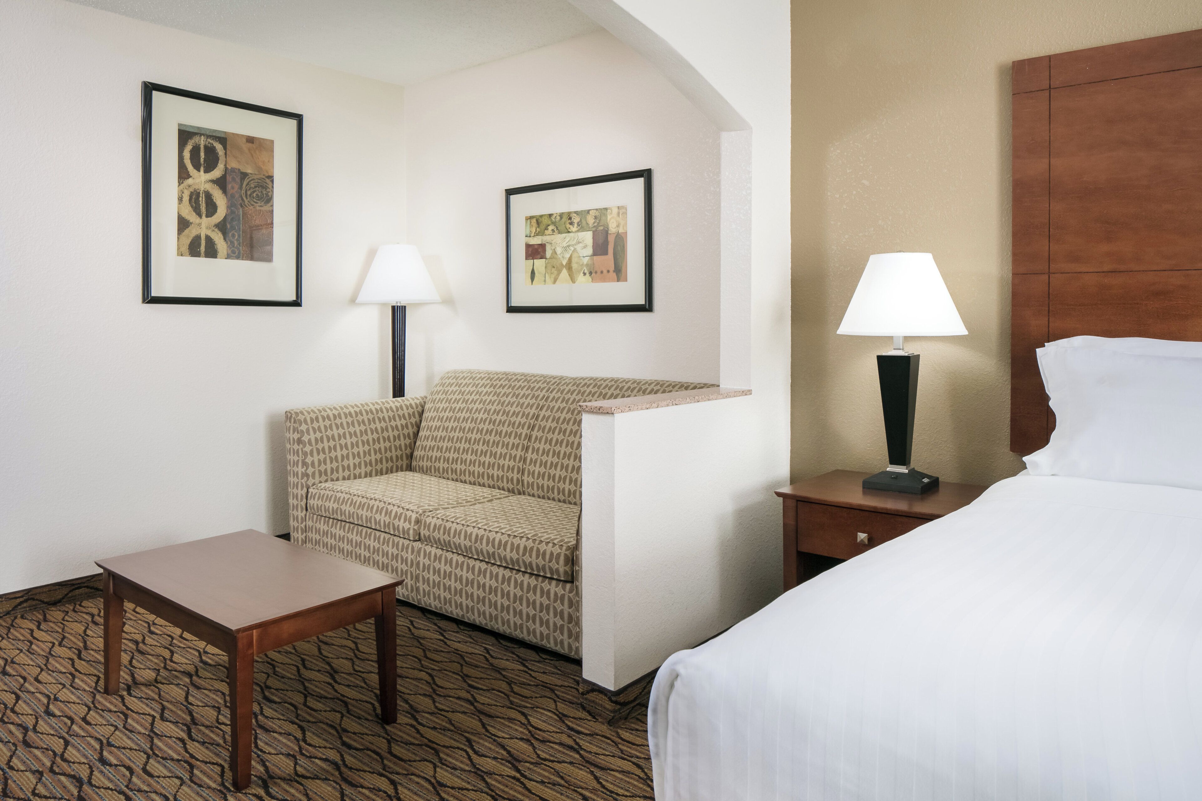 Een hotelkamer met een bed, een stoel, een tafel, twee lampen en twee foto's in lijsten aan de muur.