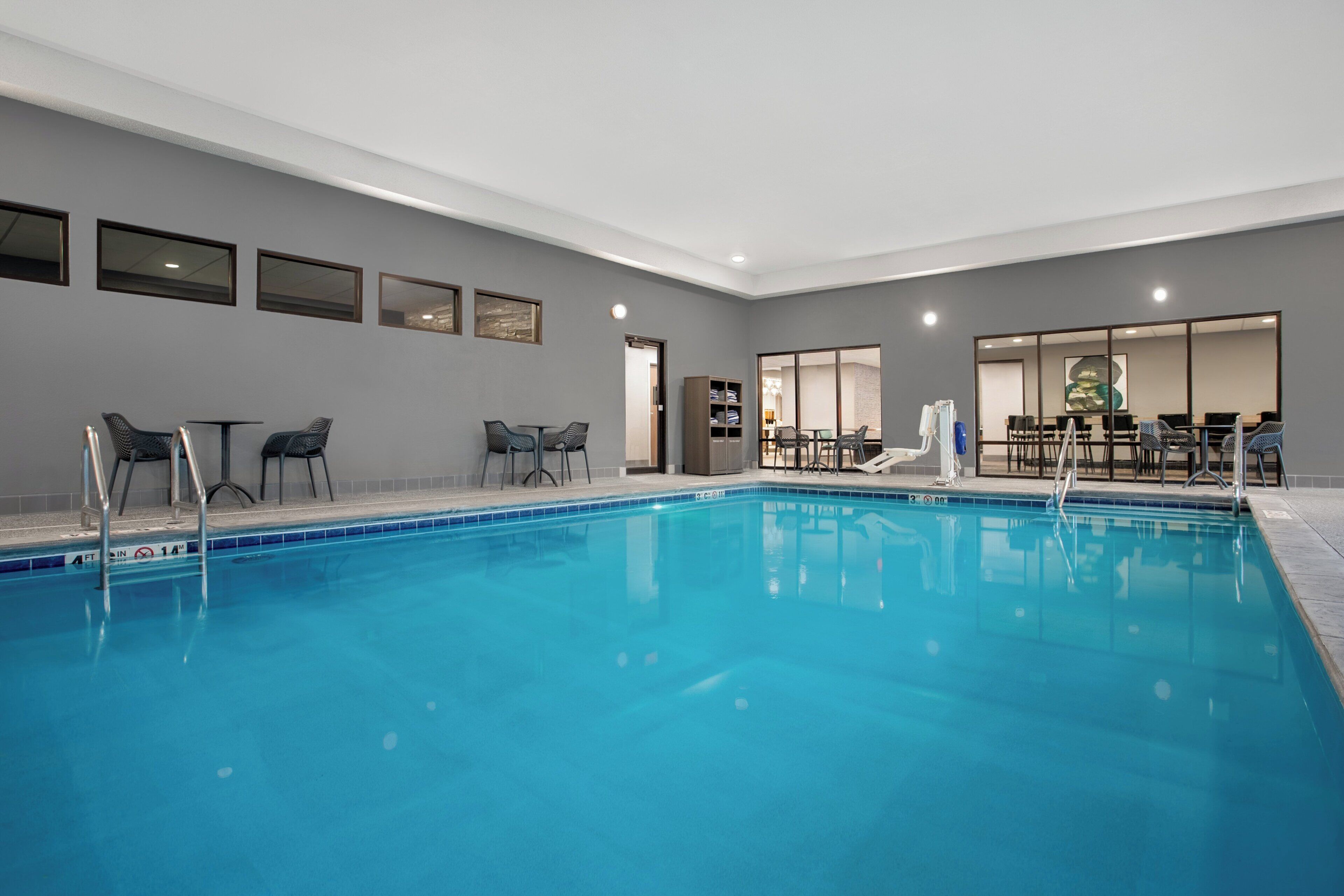A swimming pool with lounge chairs and a table, surrounded by a neutral-toned room with framed artwork on the walls.