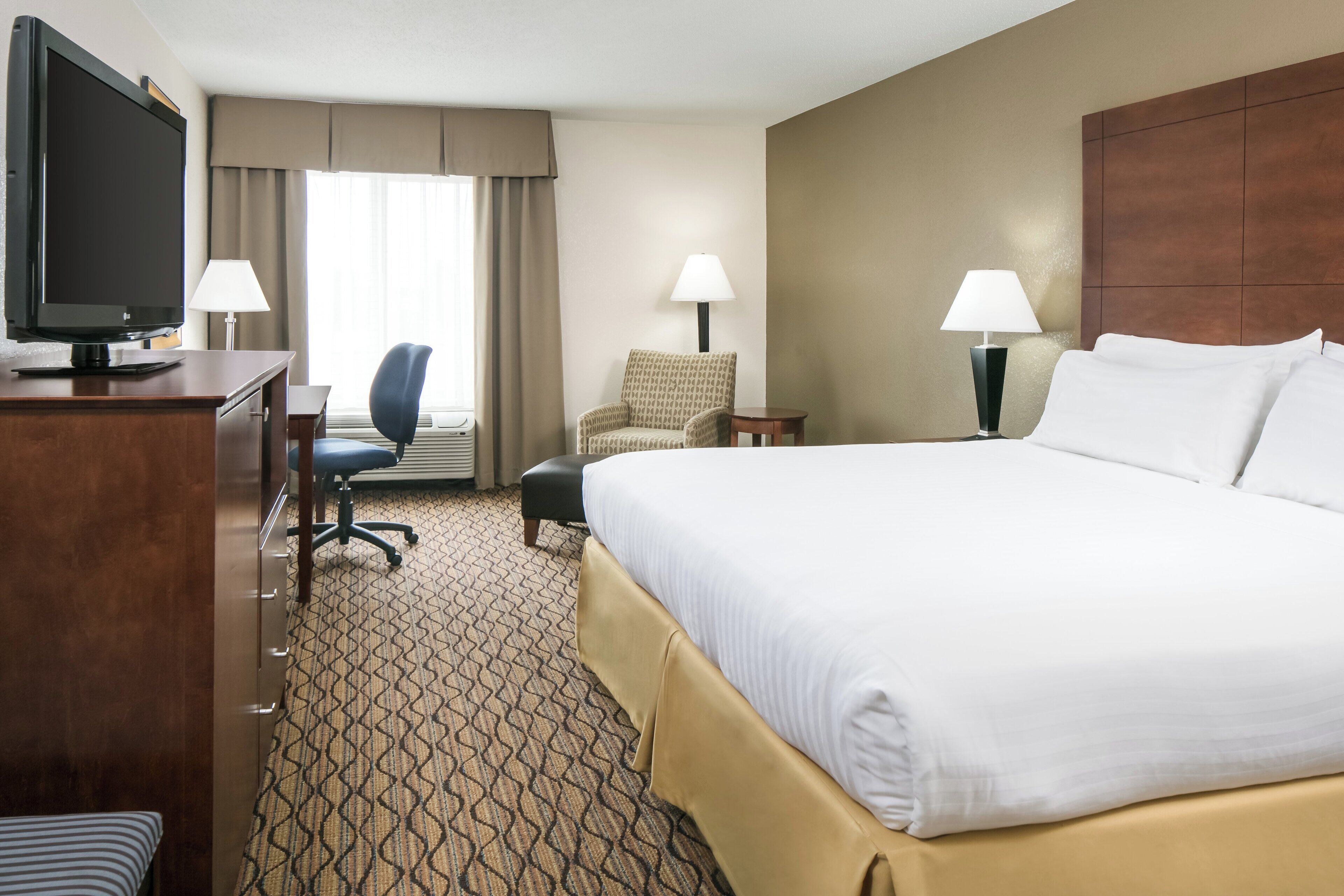 A hotel room with a large bed, a desk with a television, two chairs, a lamp, and a patterned carpet.