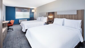 A hotel room with two beds, a desk, an orange chair, a lamp, and a window with blinds.