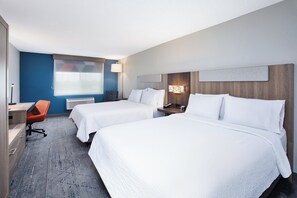 A hotel room with two beds, a desk, an orange chair, a lamp, and a window with blinds.