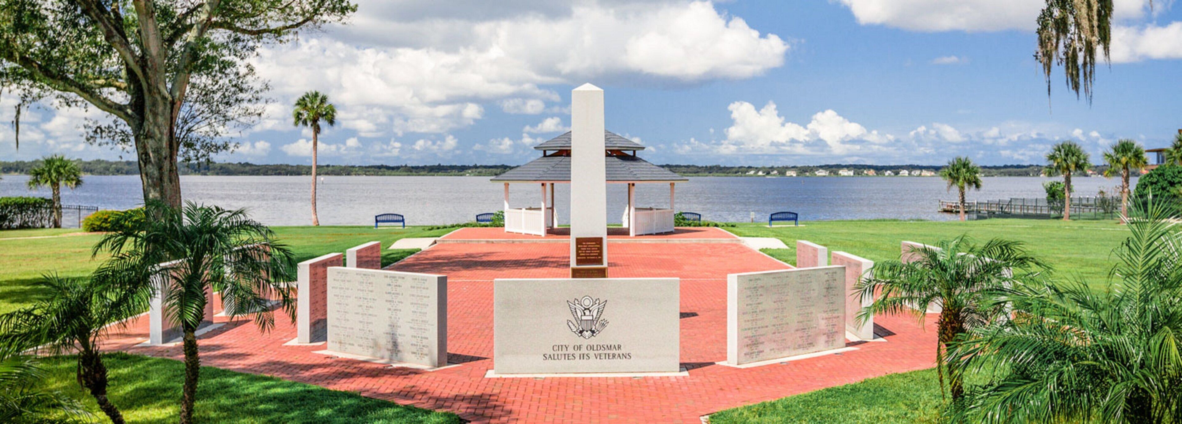 Ein Denkmal mit einem zentralen Monument und umgebenden Tafeln, vor einer Kulisse aus See und Palmen.