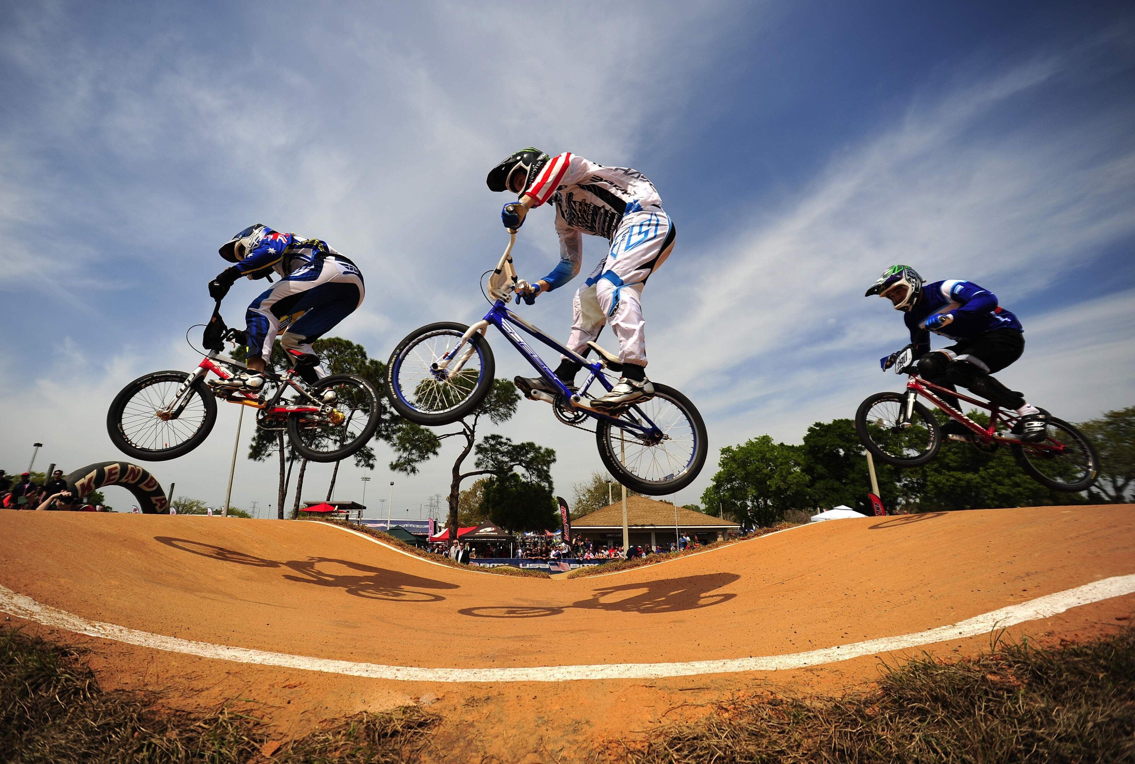 Un cascadeur de BMX effectuant une figure acrobatique sur une piste de terre, avec des spectateurs en arrière-plan.