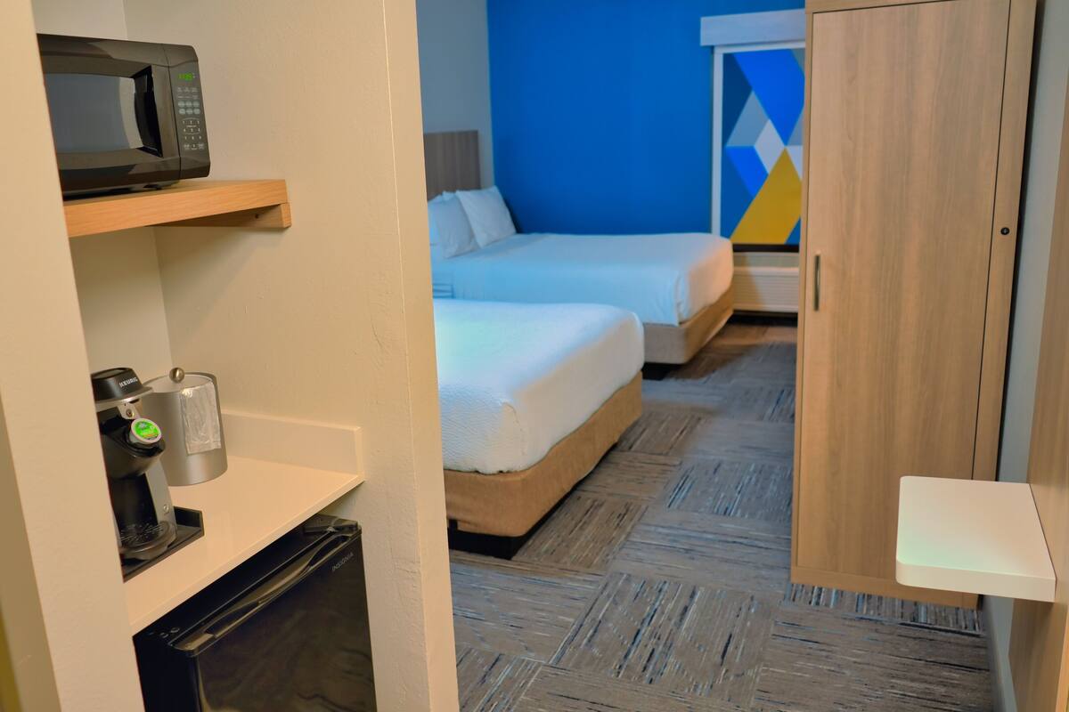 Habitación de hotel con microondas, cafetera y un refrigerador pequeño. Hay dos camas con ropa blanca y una pared azul de fondo.