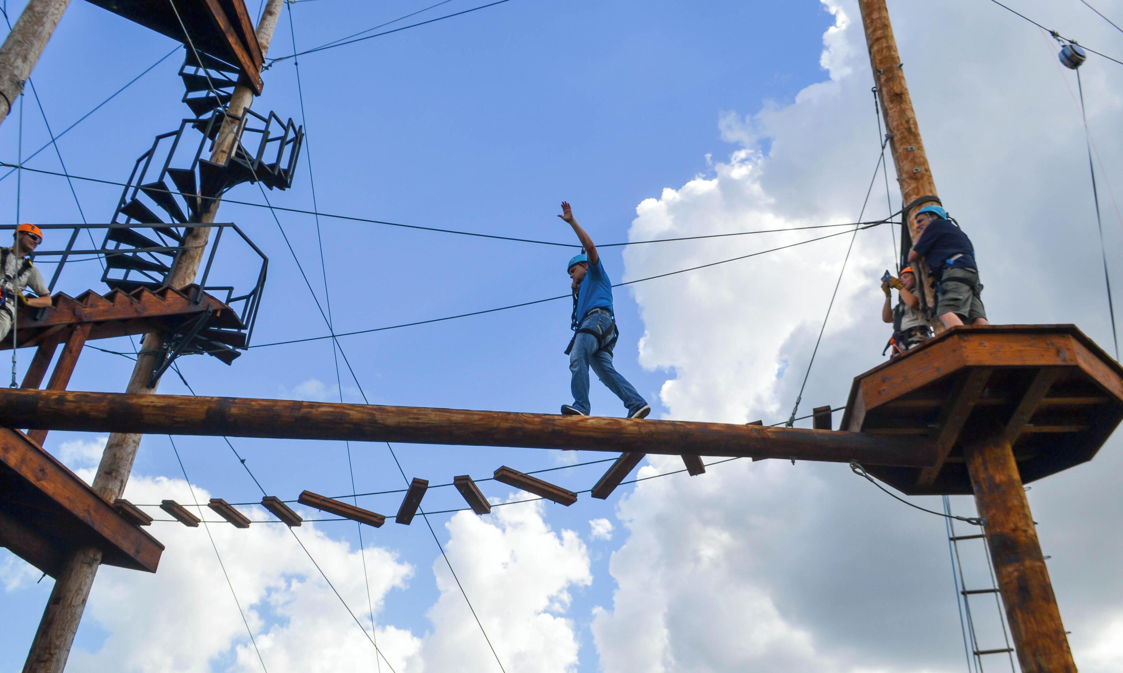 A person walking on a wooden beam structure with metal railings and cables, under a clear sky with clouds.