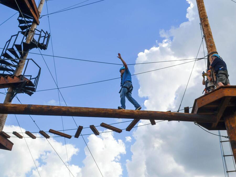 Eine Person geht über eine Holzkonstruktion mit Metallgeländern und Kabeln, unter einem klaren Himmel mit Wolken.