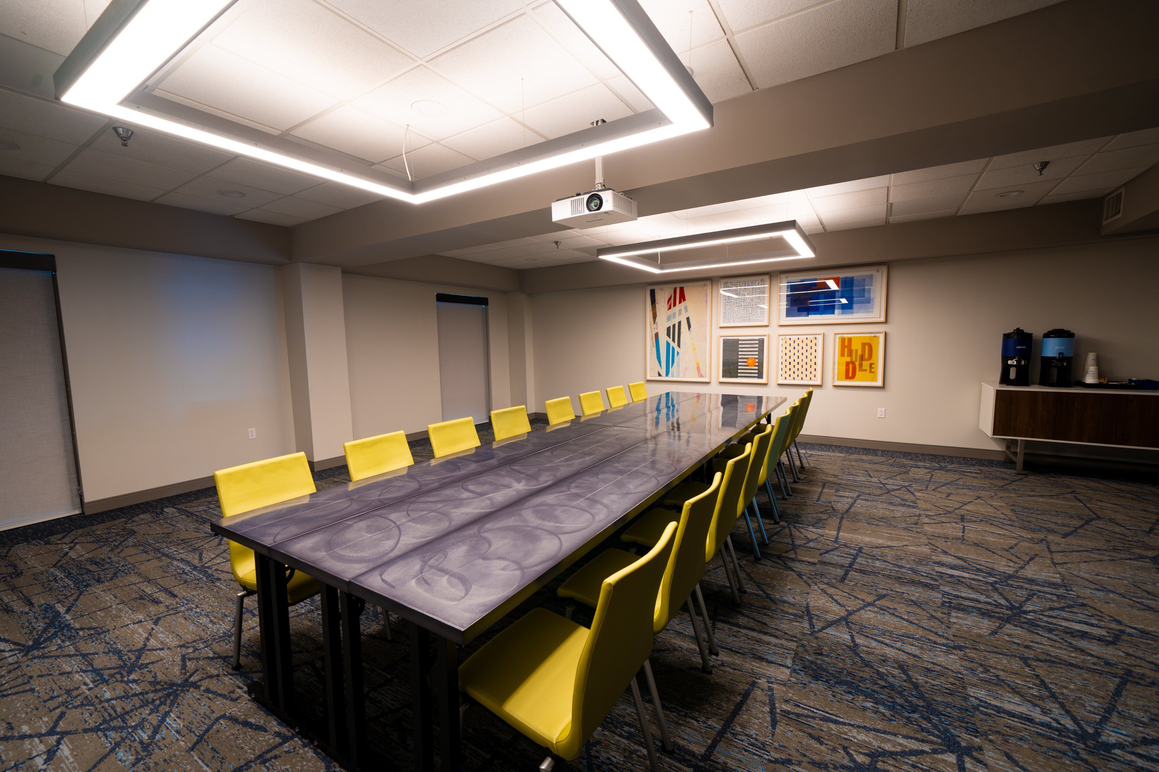 Une salle de conférence avec une longue table, des chaises jaunes, un projecteur et un sol recouvert de moquette.