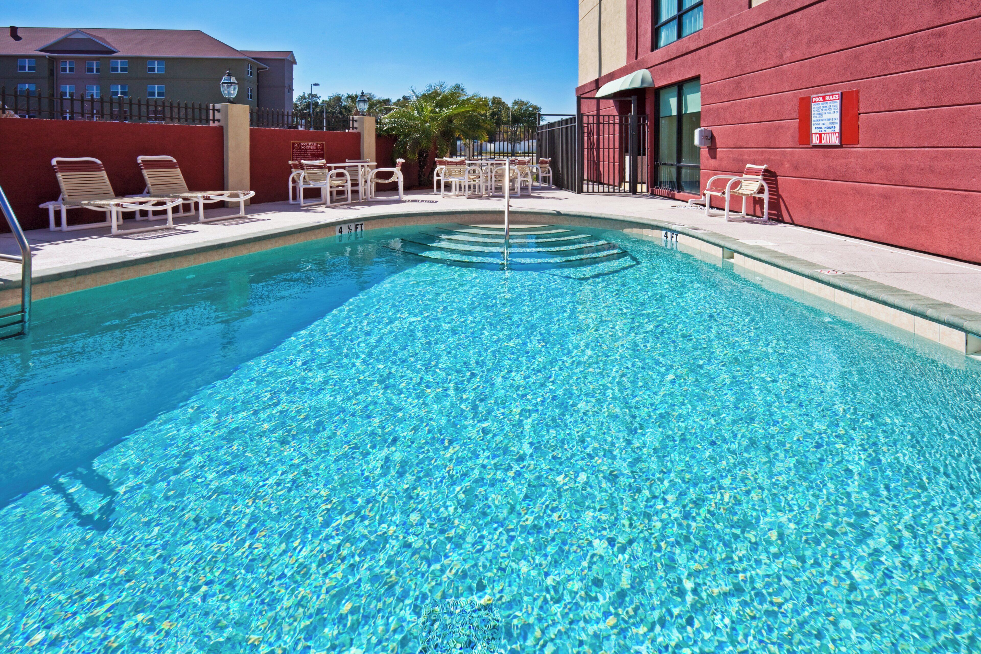 A clear blue pool with a depth marker, surrounded by lounge chairs and a red building wall.