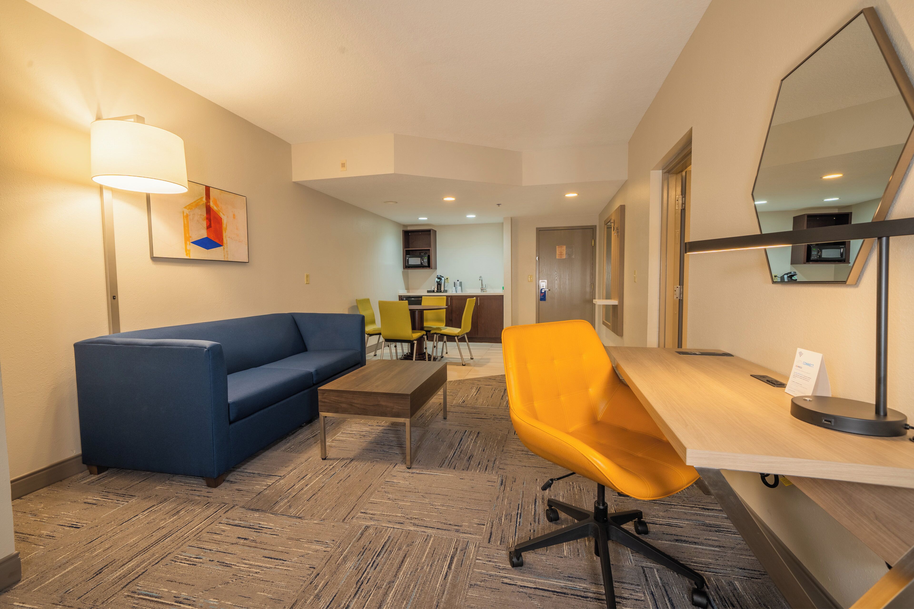 Habitación de hotel con sofá, escritorio, silla, lámpara, un cuadro y una zona de cocina al fondo.