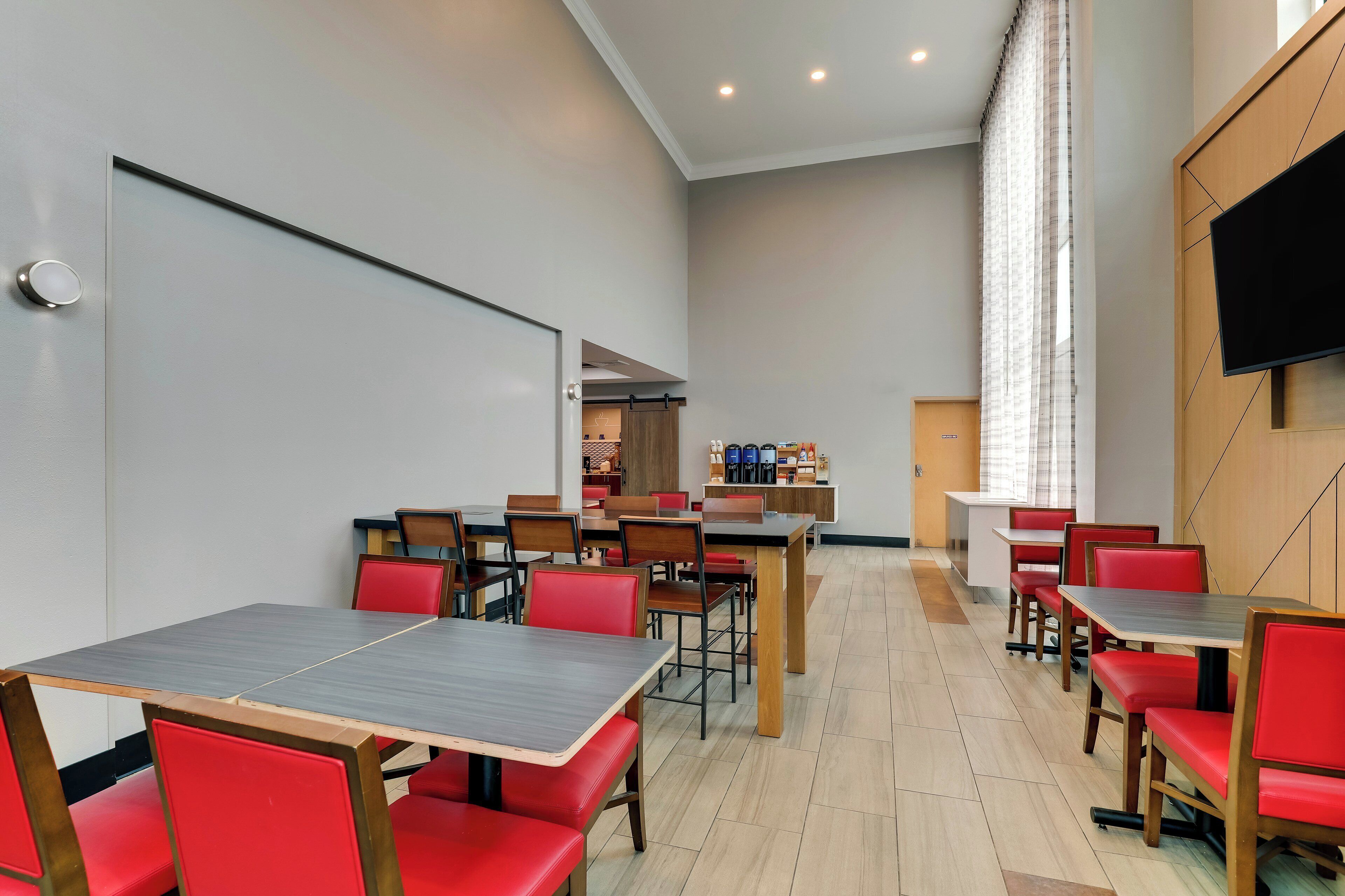 A modern dining area with multiple tables and red chairs, a flat-screen TV mounted on the wall, and a large window with curtains.